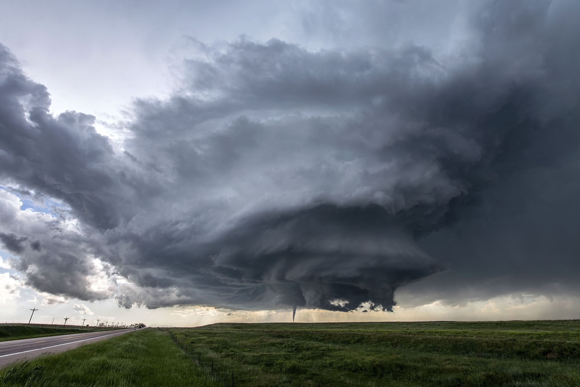 Dark storm clouds with a visible tornado extending to the ground over a green field and road.