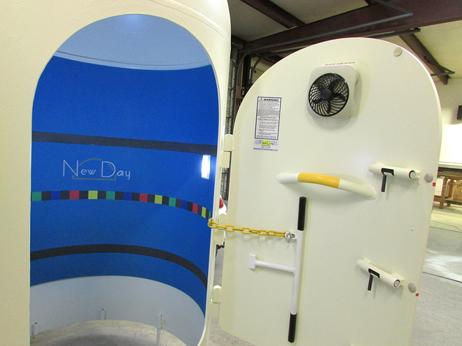 White hyperbaric chamber with open door, interior blue, fan, yellow handle, and decorative colored lines.