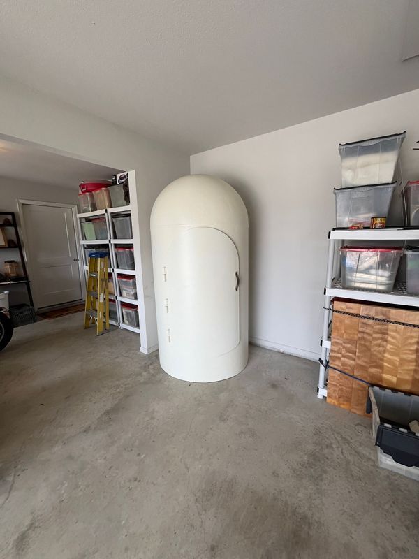 White, cylindrical storm shelter in a garage, next to shelves with storage bins.
