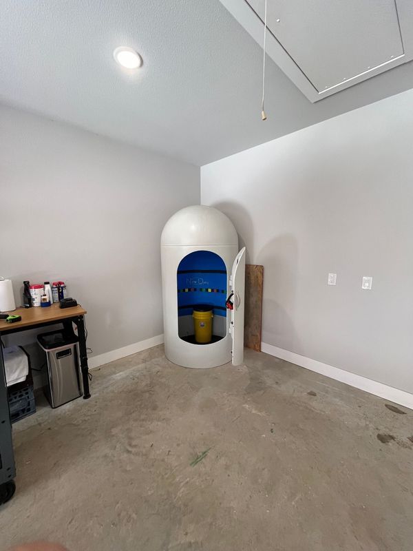 A sensory deprivation float tank in a garage. The tank is white and cylindrical with an open blue interior.