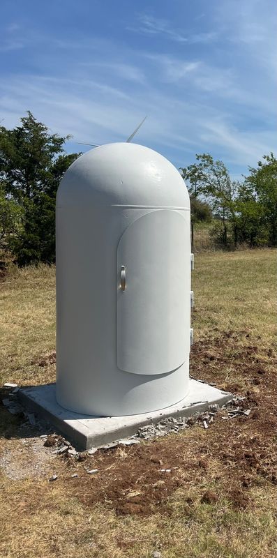 A large, gray, cylindrical outdoor equipment box with a rounded top, door, and lightning rod on a concrete base.