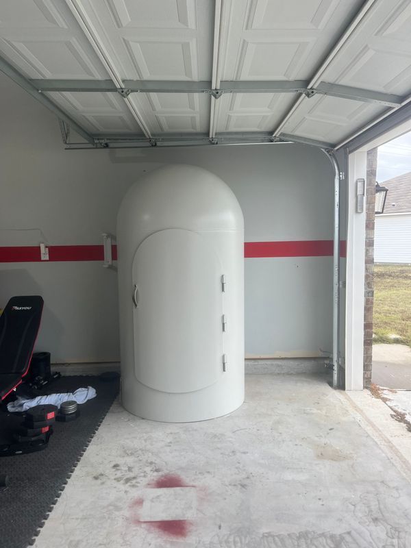White cylindrical hyperbaric chamber inside a garage. Door is closed, with three small handles. Red stripe on wall.