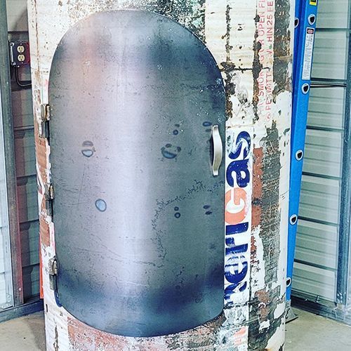 A metal door is installed on a cylindrical tank covered in peeling paper. A ladder is positioned next to the tank.