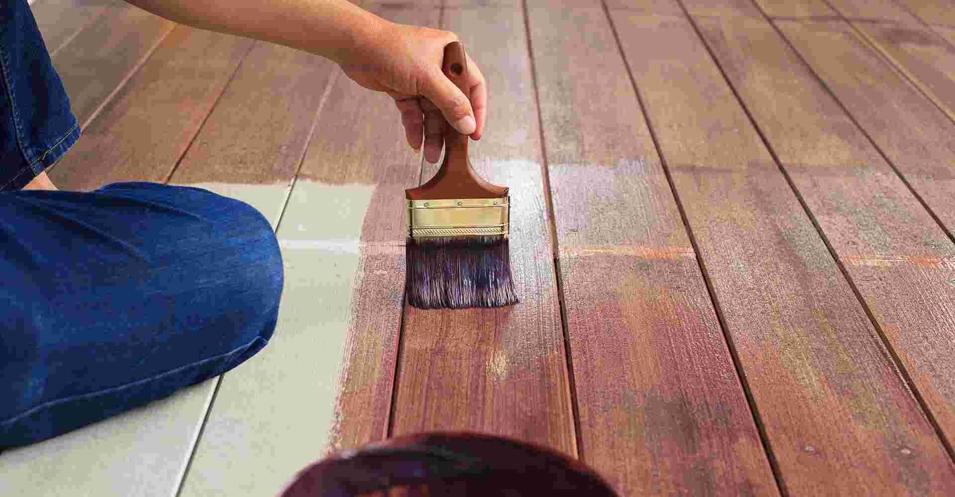Hand applying a clear sealant to wooden floorboards with a paintbrush.