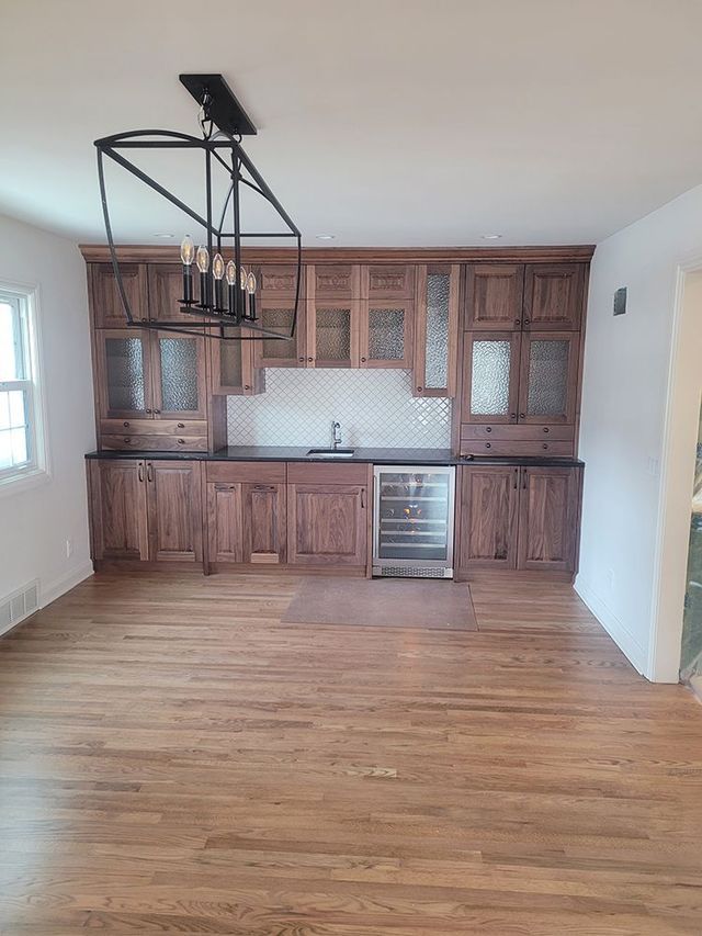 Wooden bar with wine cooler and cabinets, under a black chandelier. Light-colored backsplash. Wooden floor.