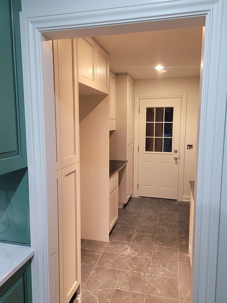Interior view of a pantry with white cabinets, a refrigerator, and a door leading outside with a gray tiled floor.