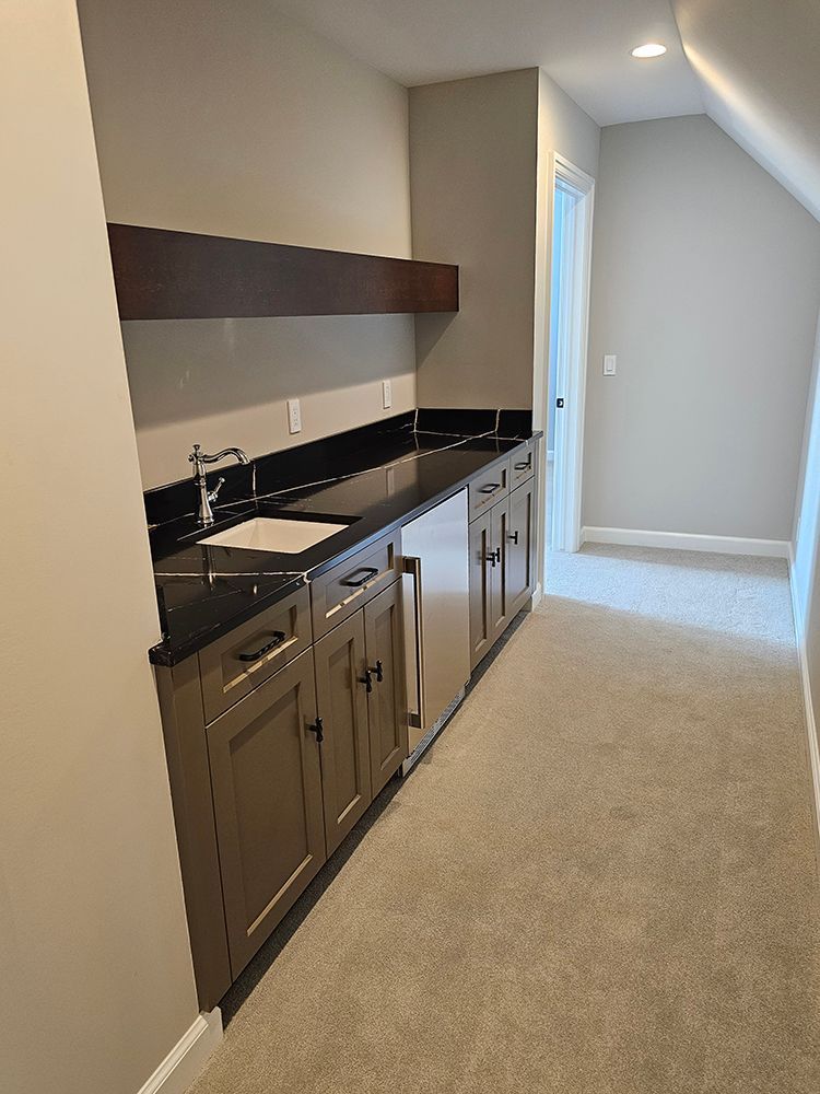 Small wet bar with sink, cabinets, countertop, and refrigerator in a hallway, tan and brown tones.