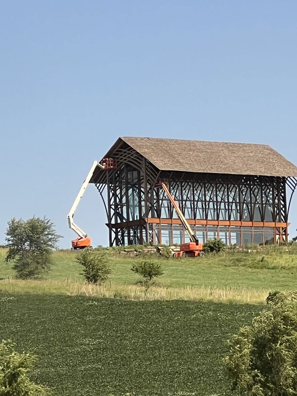 Barn under construction on a grassy hill; two orange lifts work on the facade, against a blue sky.