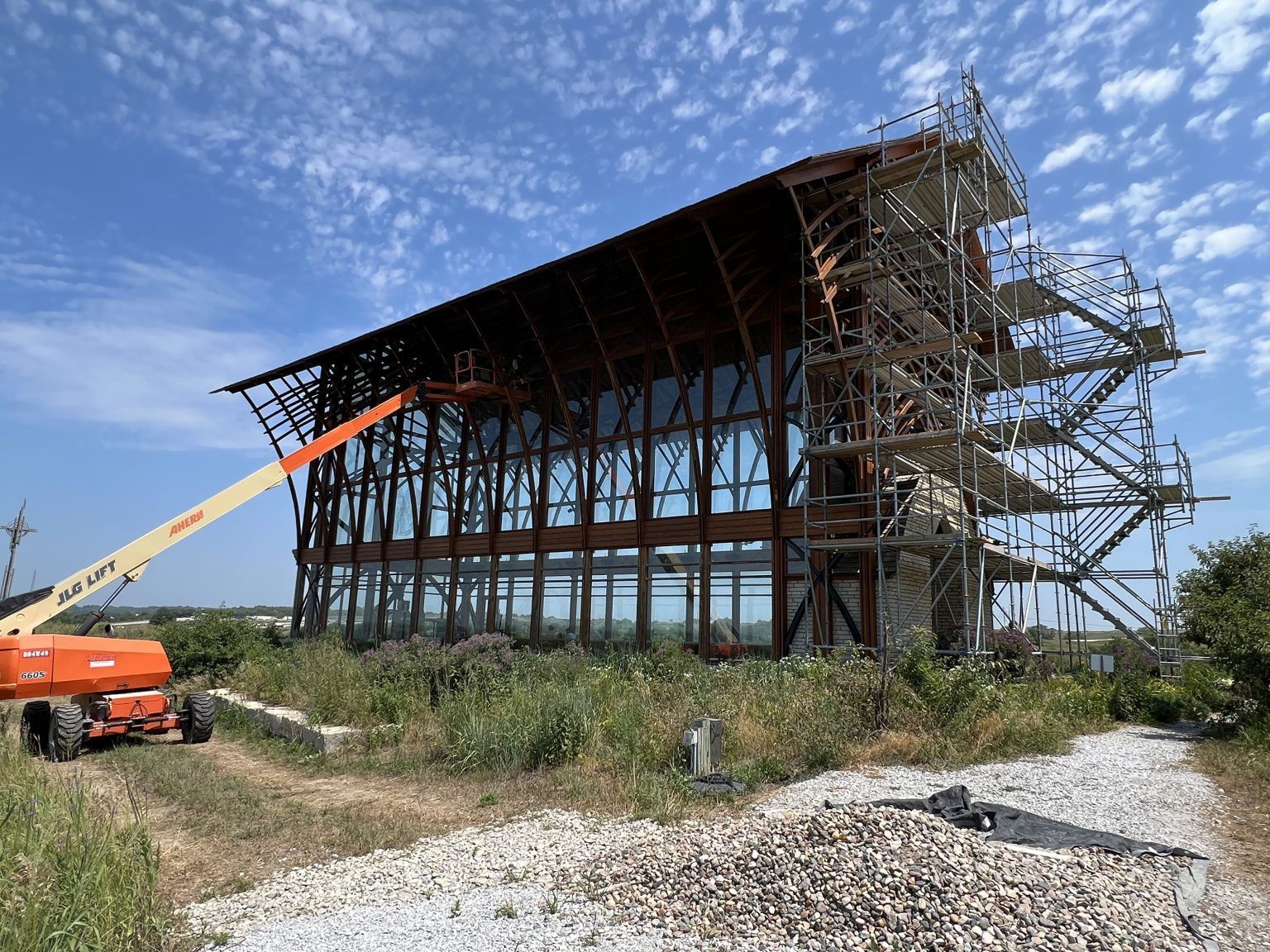 Building under construction with scaffolding and an orange lift on a gravel path under a blue sky.