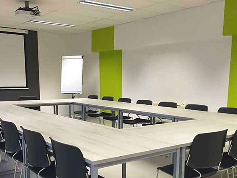 Empty conference room with square table and chairs, projection screen, and flip chart. Lime green accents.