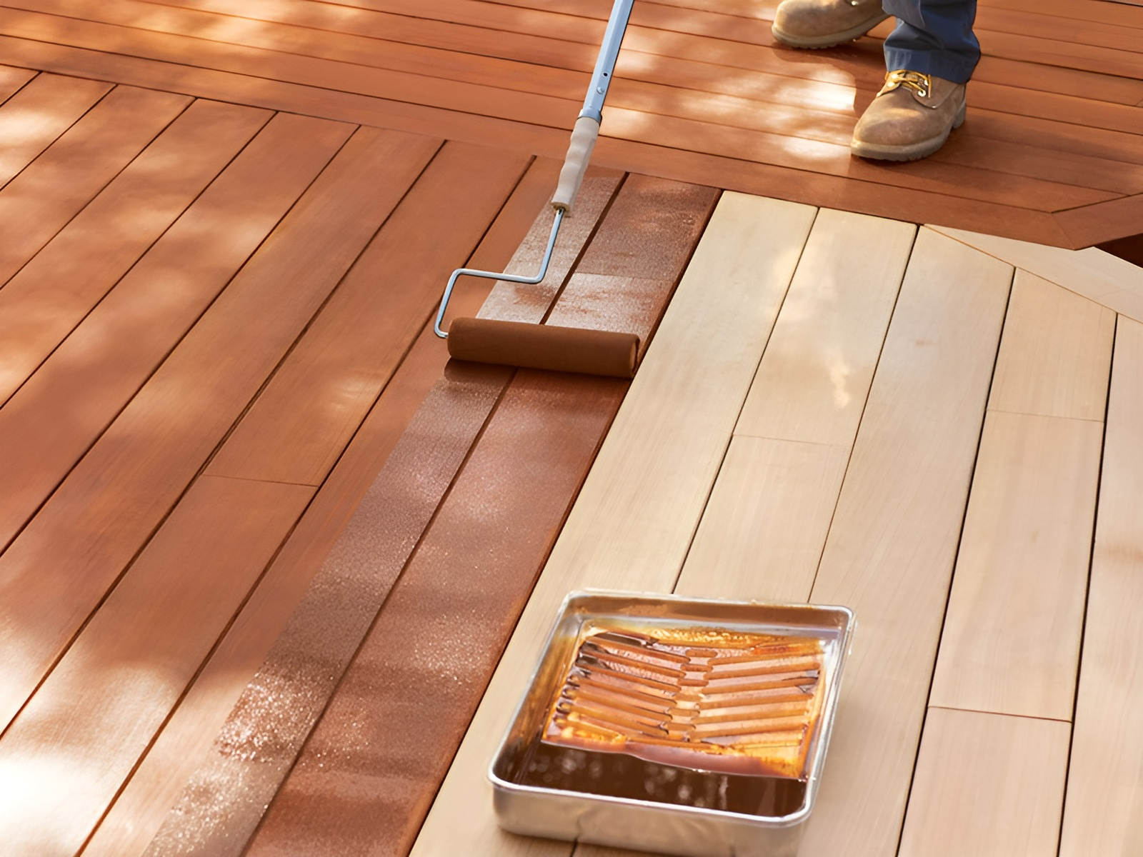 Person applying stain to a wooden deck with a roller. Tan stain, brown deck boards, outdoor setting.