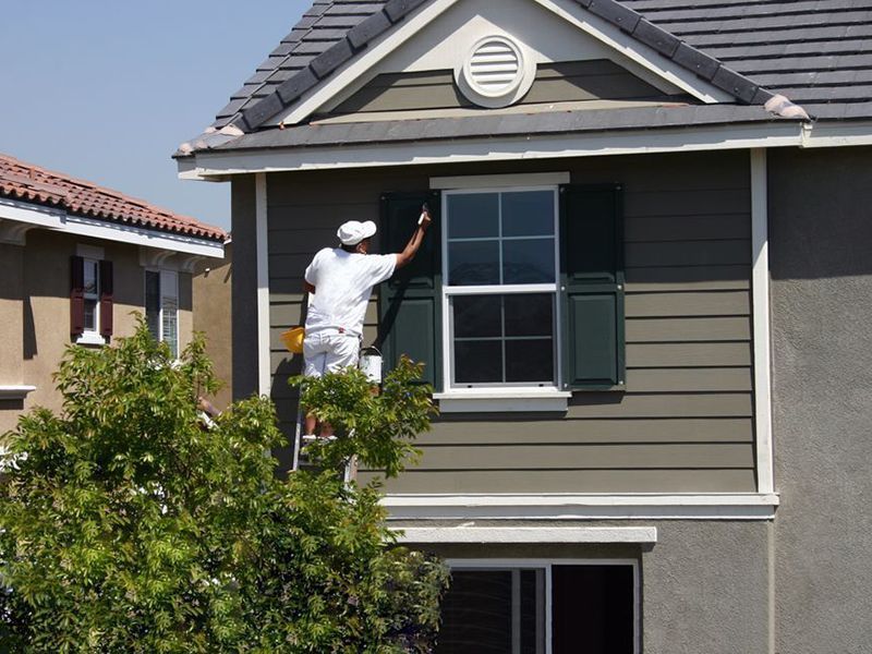 Painter on a ladder painting a house's shutters green, exterior view.