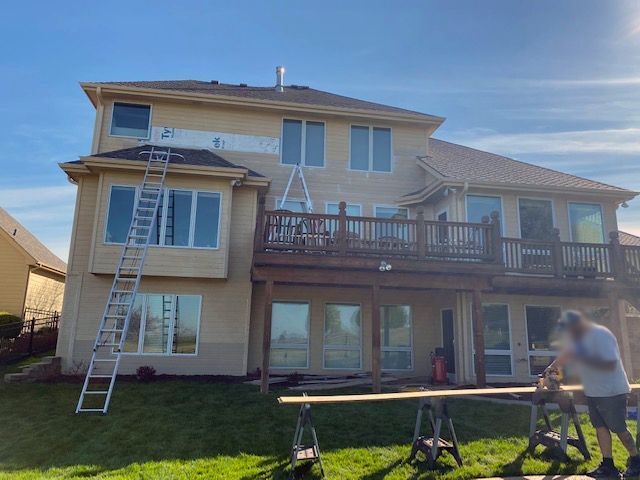 House exterior being worked on, ladders, scaffolding, worker cutting wood, blue sky, green grass.