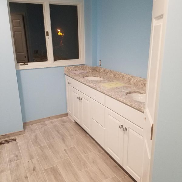 Bathroom with blue walls, white cabinets, granite countertop, two sinks, and wood-look tile flooring.