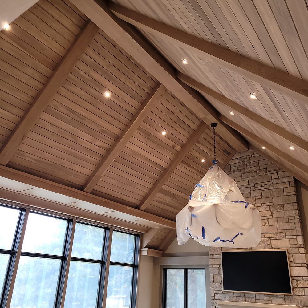 Vaulted wood ceiling with recessed lights and a light fixture, over a living space with windows and a stone fireplace.