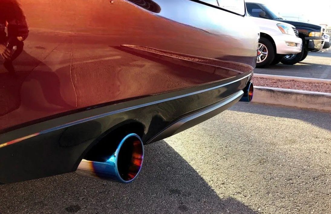 Close-up of a dark red car's bumper and a custom, burnt-titanium-style exhaust tip in a parking lot.