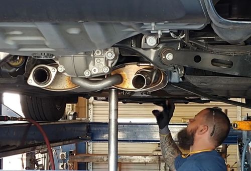 A mechanic works under a car in a repair shop, using a tall pole jack to support the exhaust system.