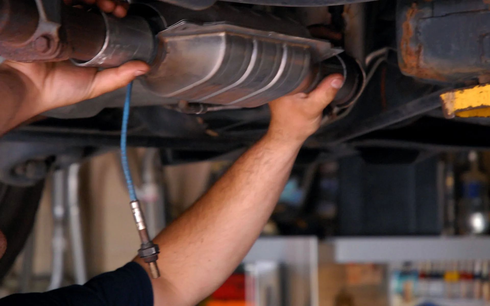 A mechanic works under a car to install or replace a catalytic converter, holding the metal component in place.