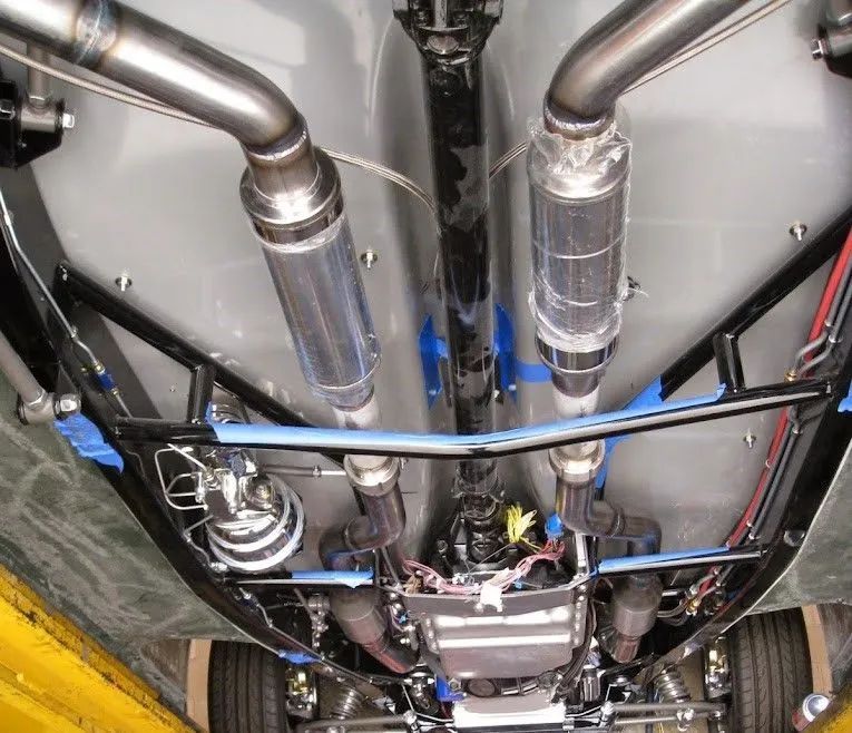 An underside view of a vehicle showing a dual exhaust system, custom black chassis frame, and transmission components.
