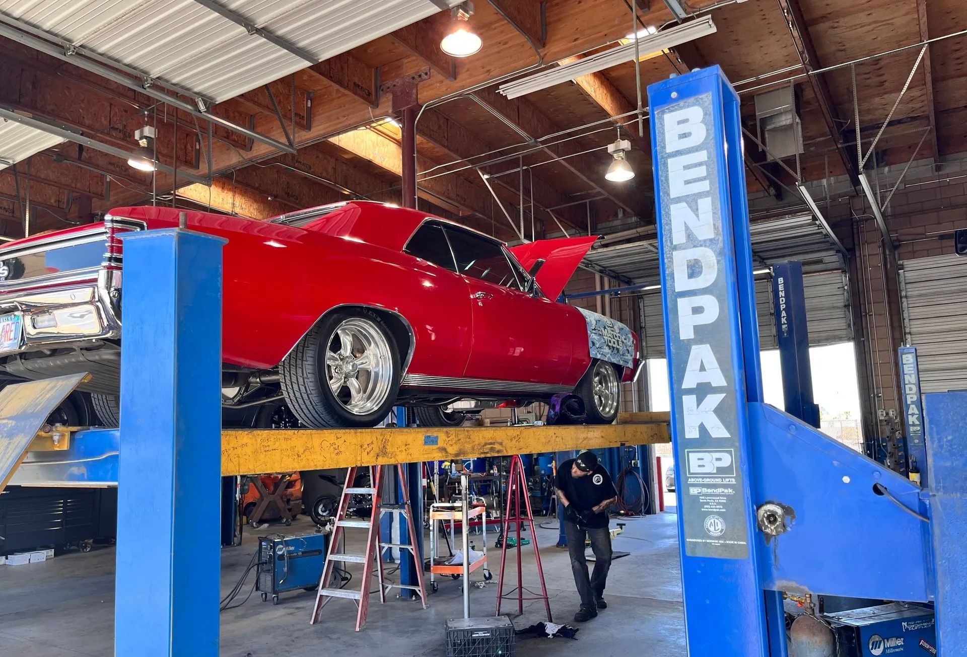 A red muscle car is elevated on a blue BendPak lift in a garage, while a person works underneath it.