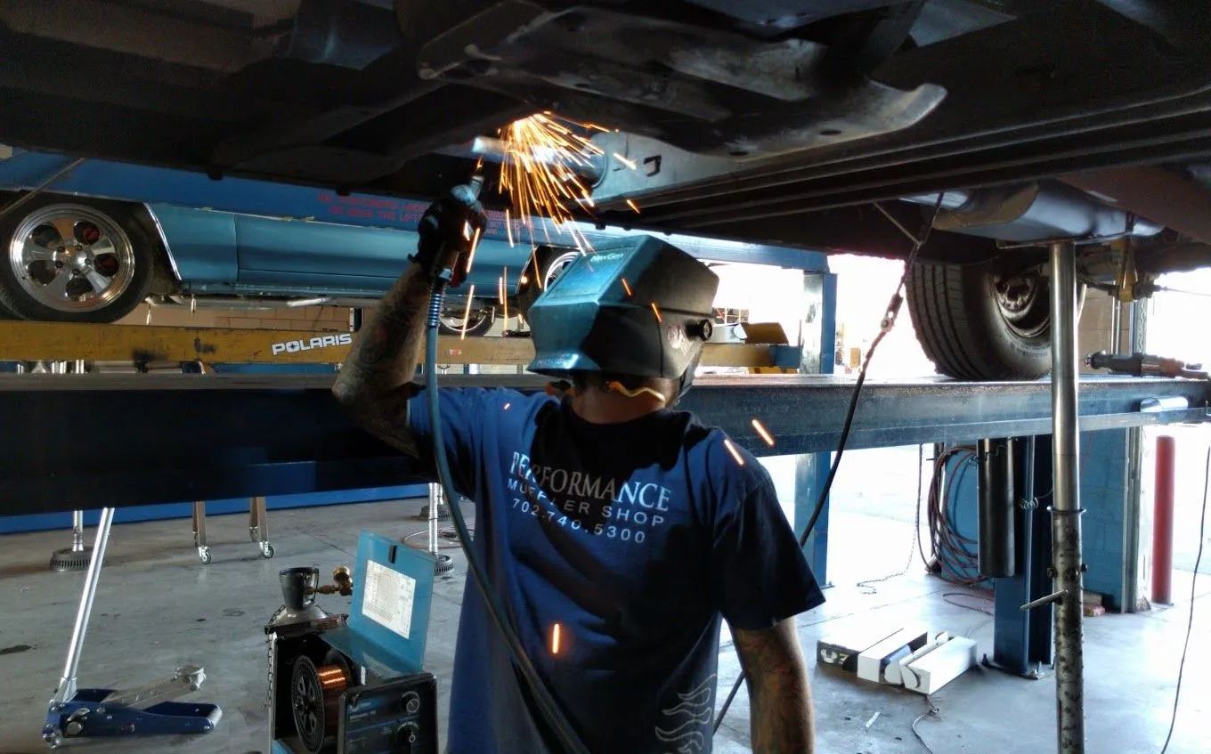 A worker in a blue shirt and protective helmet grinds metal beneath a vehicle lifted in an auto repair shop.