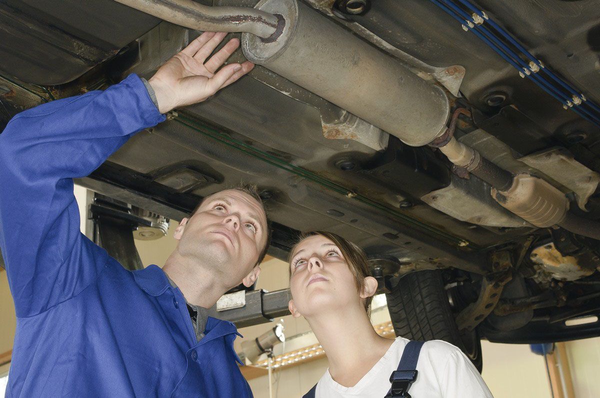 A mechanic and an apprentice looking up at the undercarriage of a car raised on a lift in a workshop.