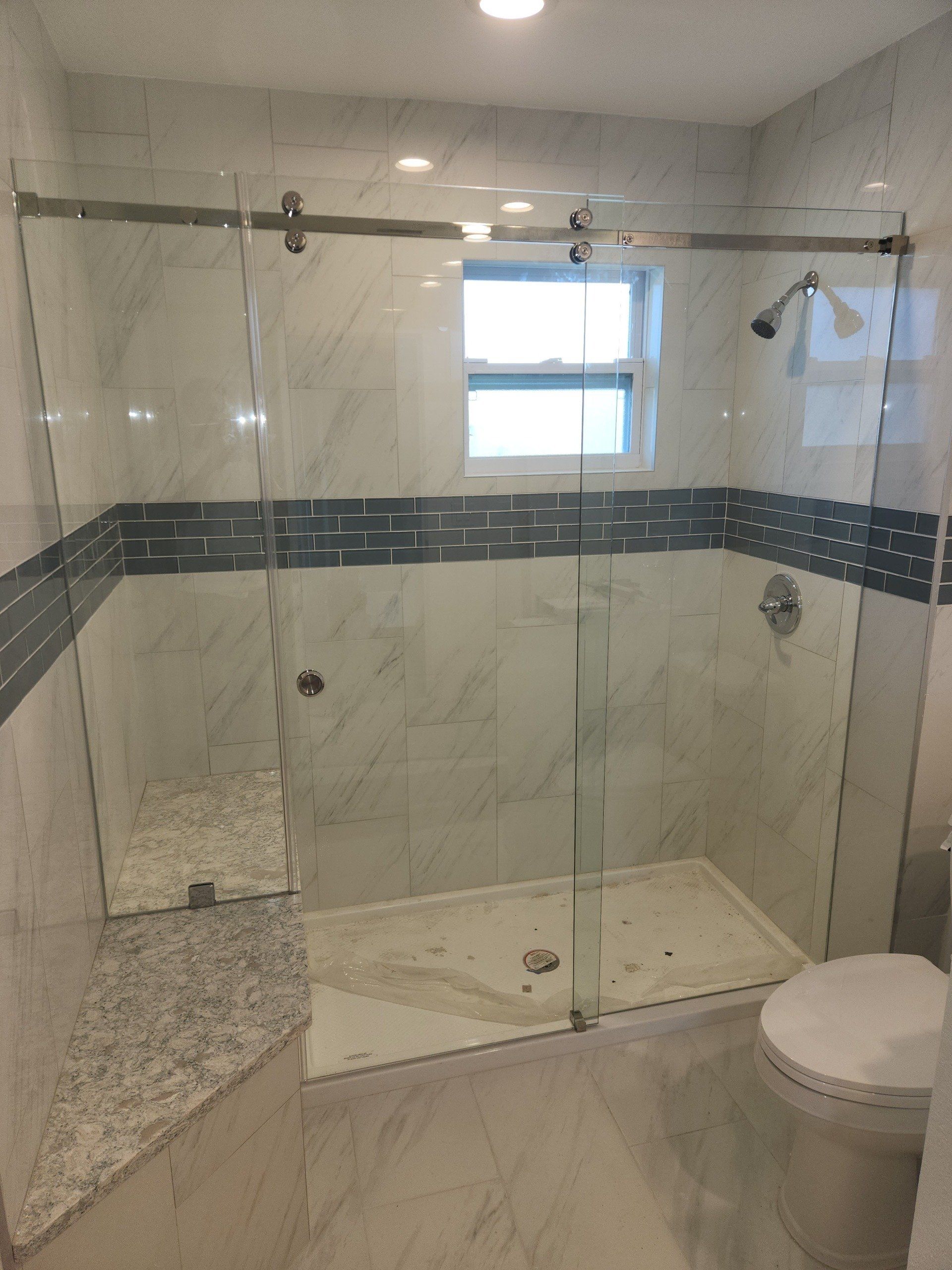 Glass shower with sliding door, white marble tile, gray accent, toilet, and window in a bathroom.