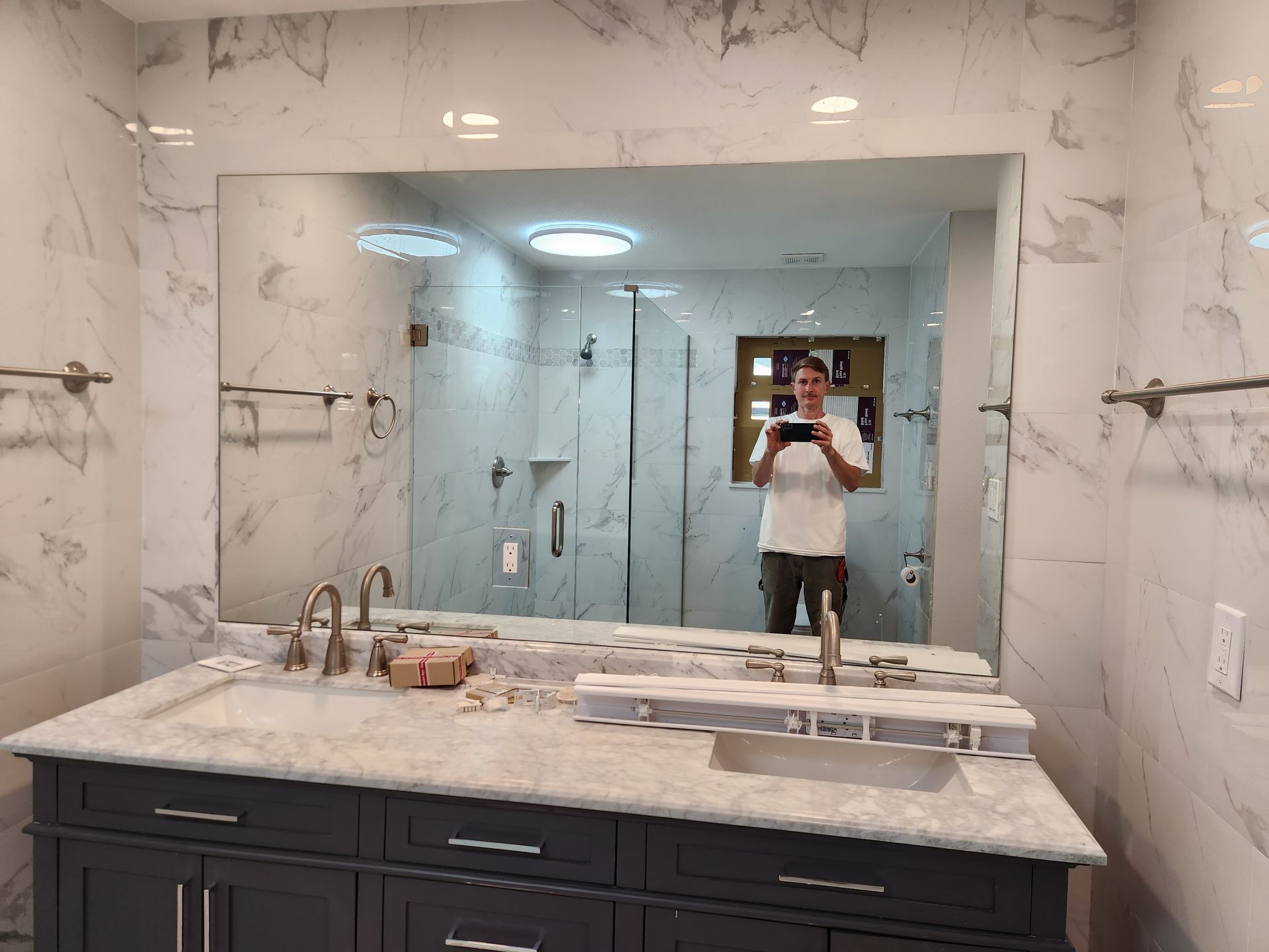 Person taking a photo in a modern bathroom with marble walls, a double sink vanity, and a large mirror.