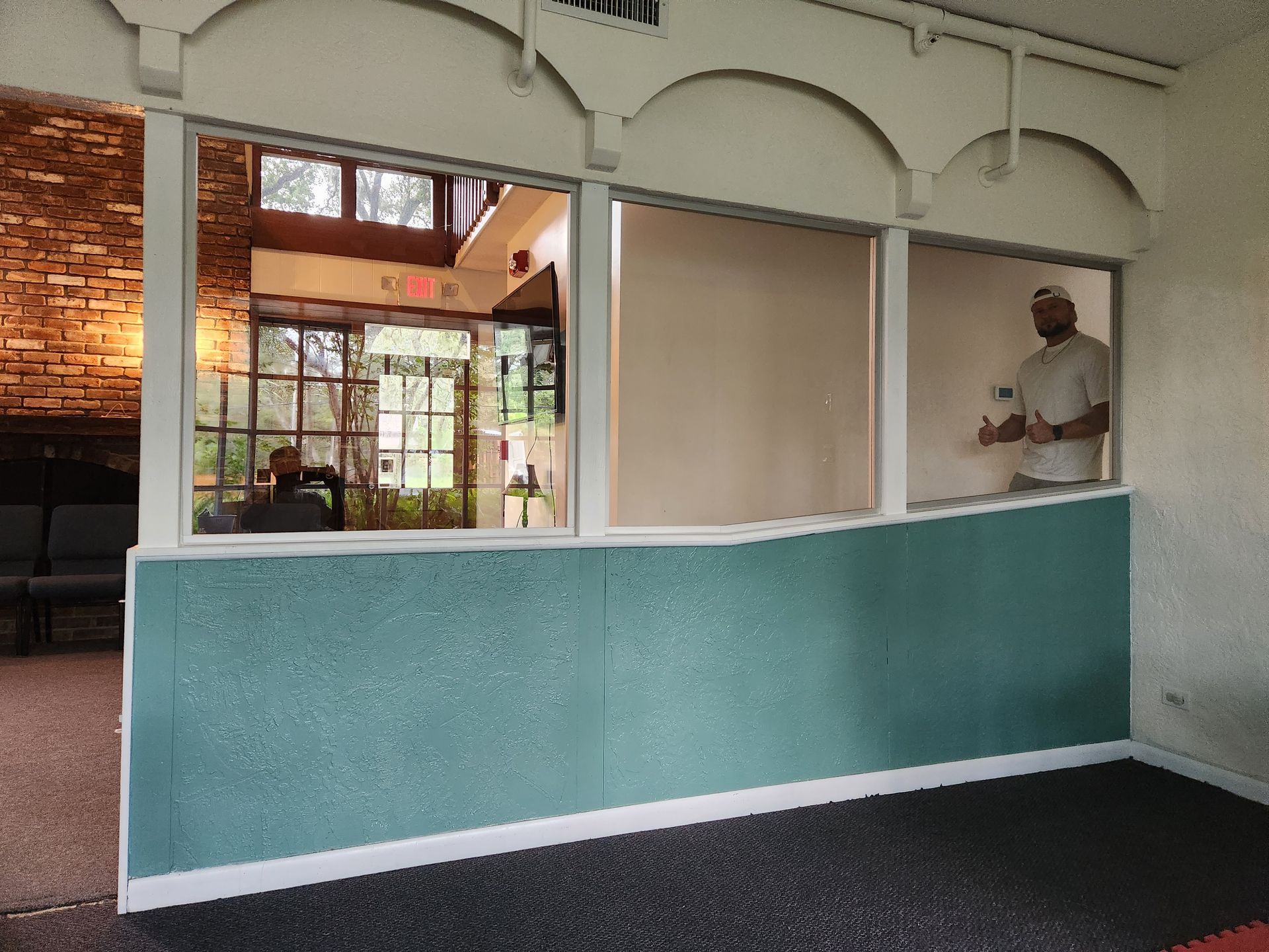 A person standing by a window with a teal wall. Inside, an older man sits near a brick wall.