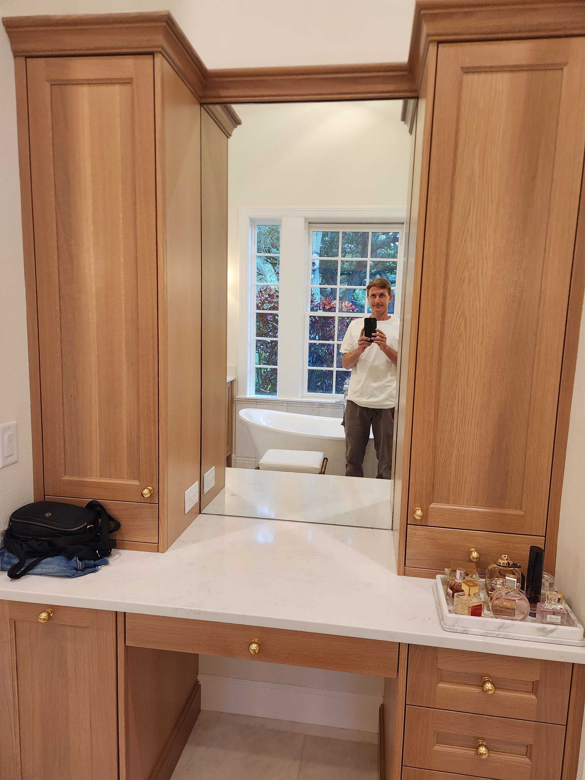 Man in a bathroom mirror, standing at a vanity with light wood cabinets, a white countertop, and a window.