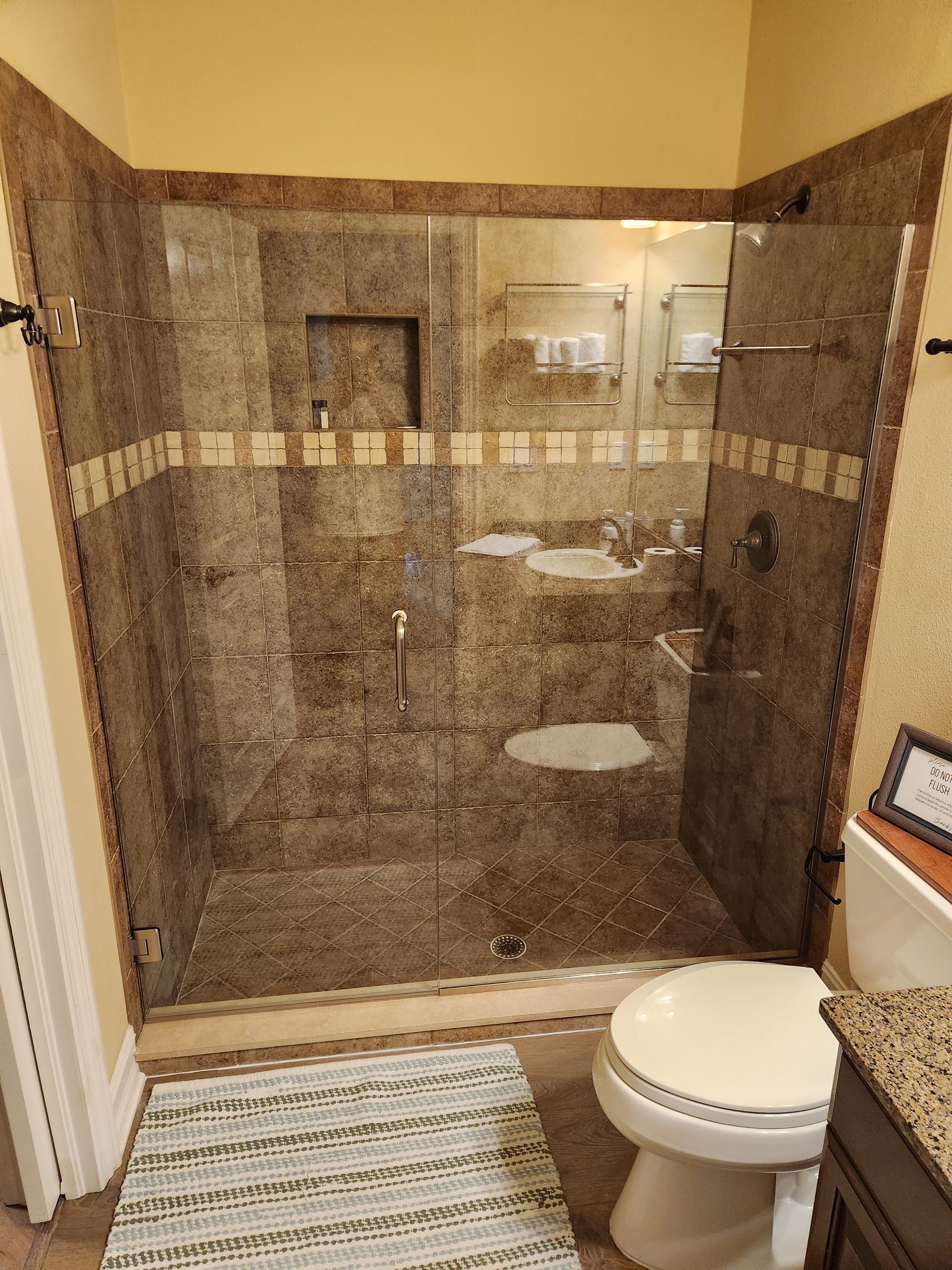 Bathroom with a brown tile shower, a white toilet, and a small rug.