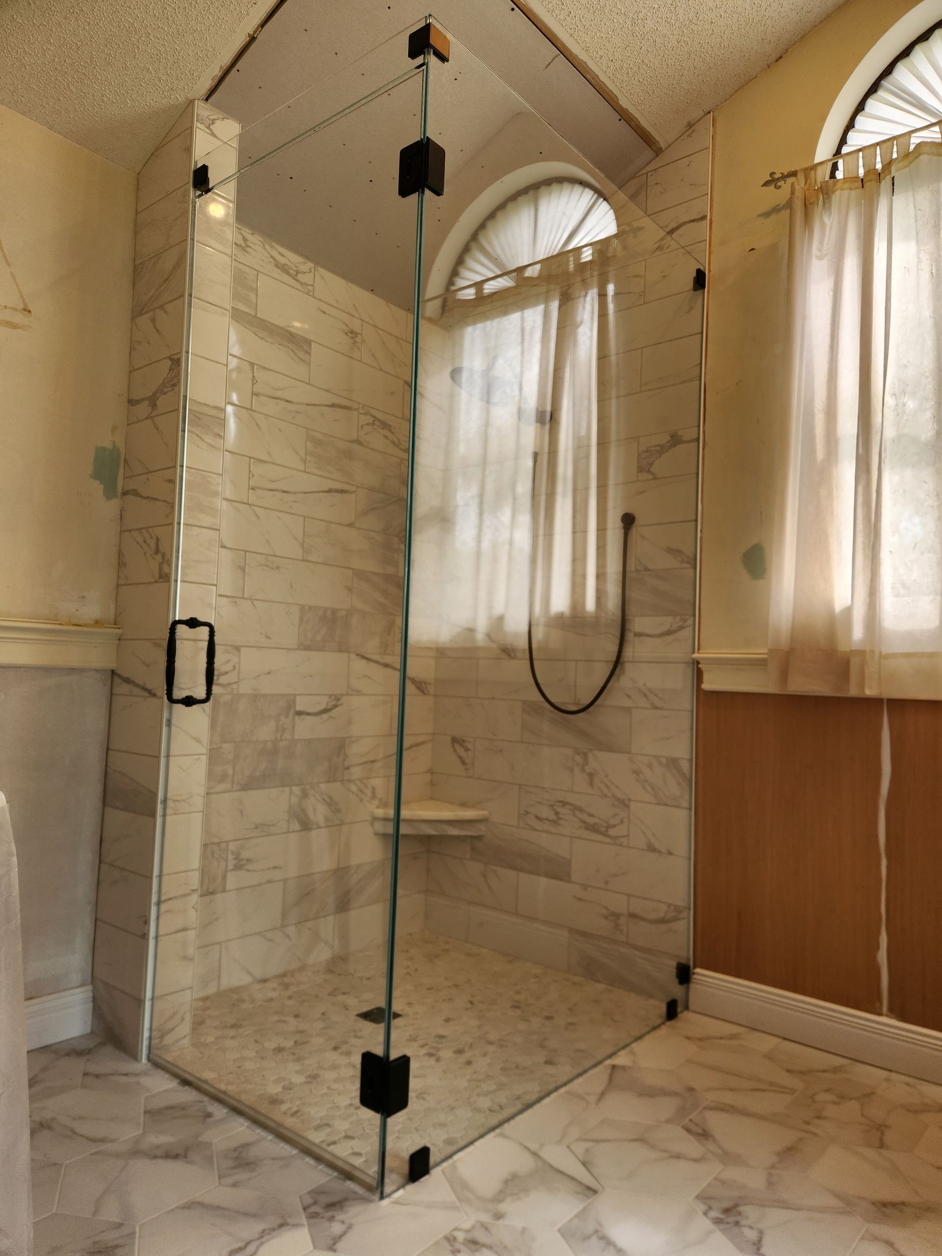 Glass shower enclosure in a bathroom with marble-look tiles, next to a window with curtains.