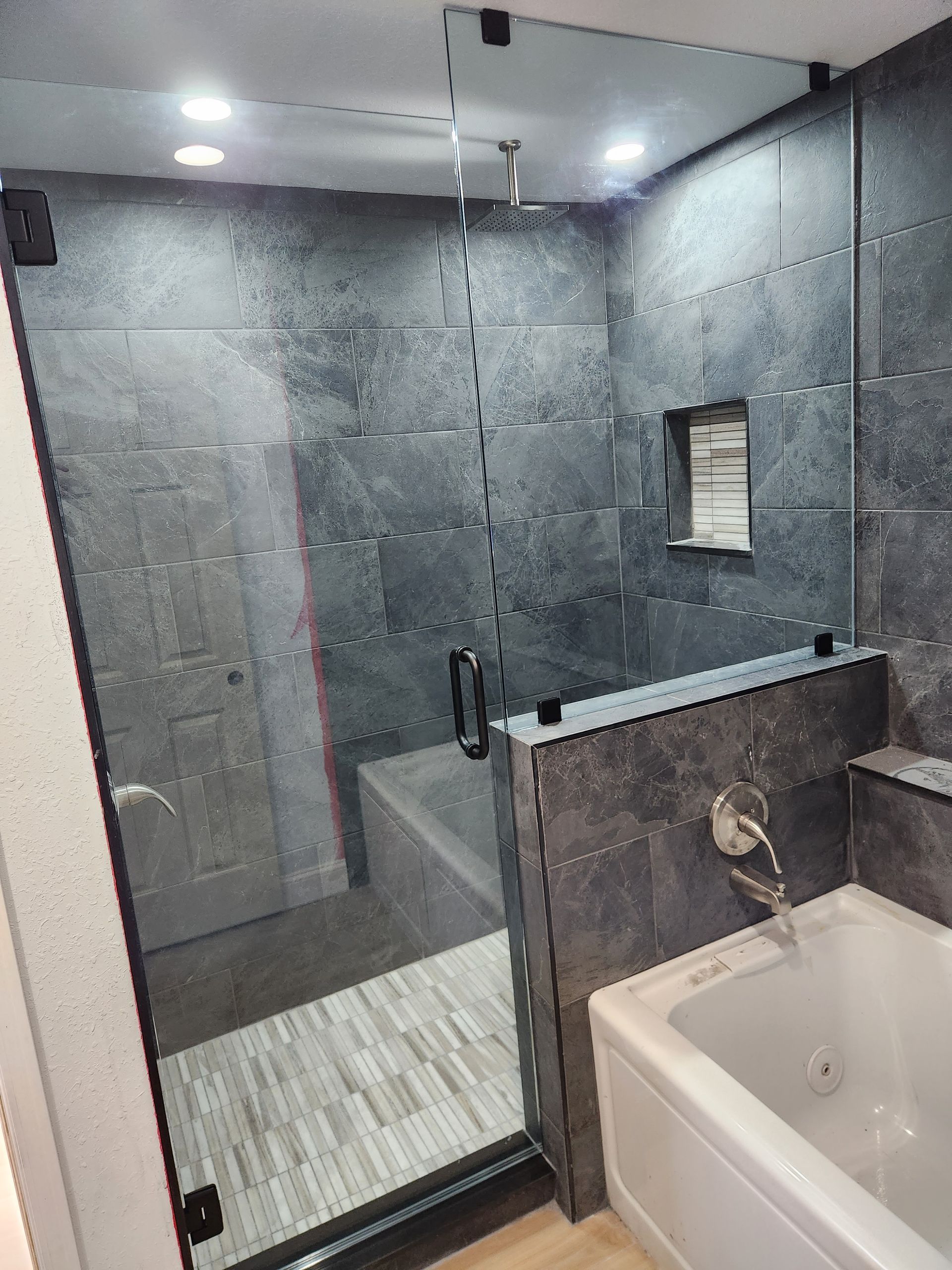 Modern bathroom with a glass shower door, dark tile, and a white bathtub.