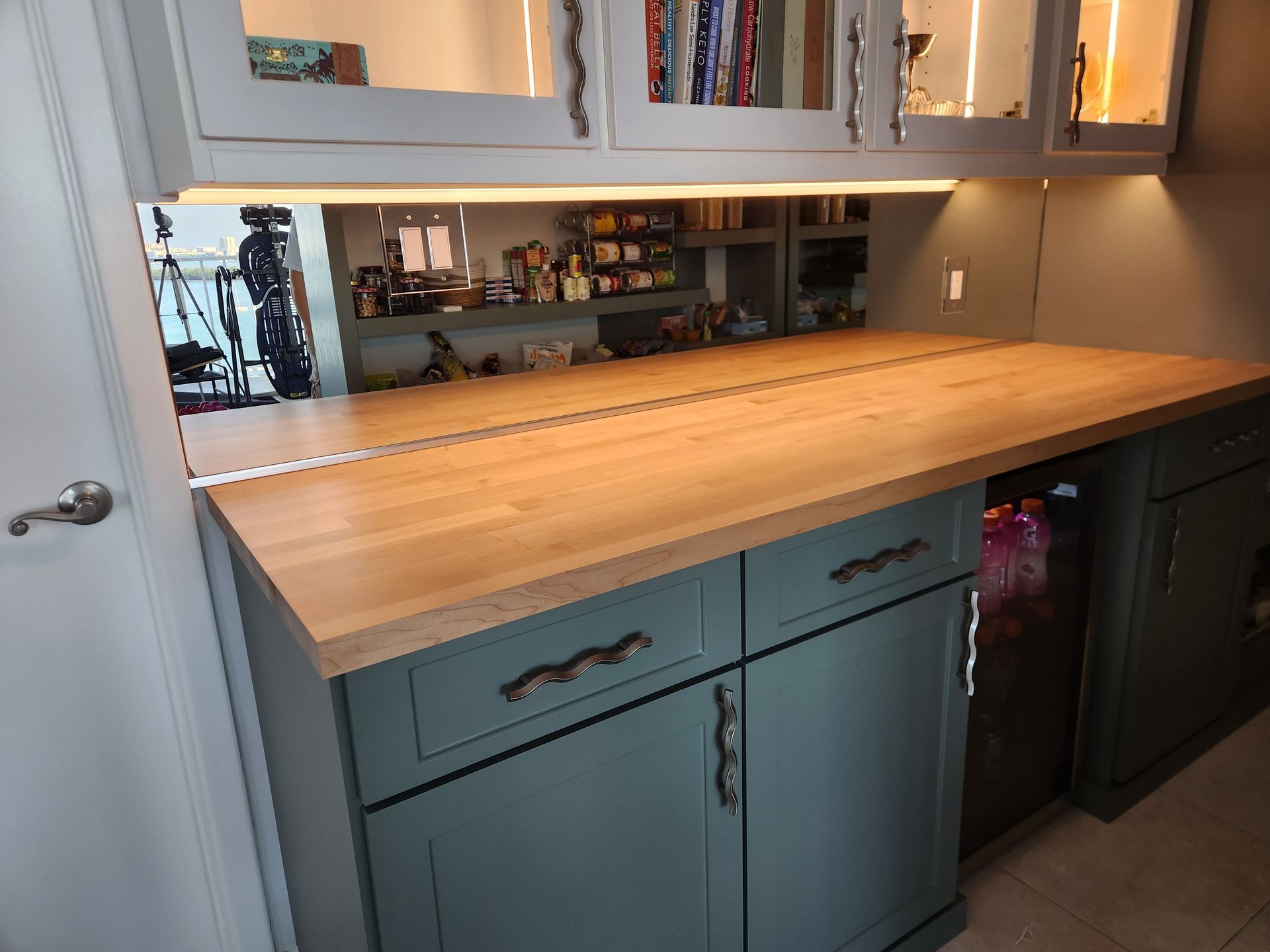 Wooden countertop on green cabinets with a built-in wine fridge, under-cabinet lighting, and glass-front cabinets above.