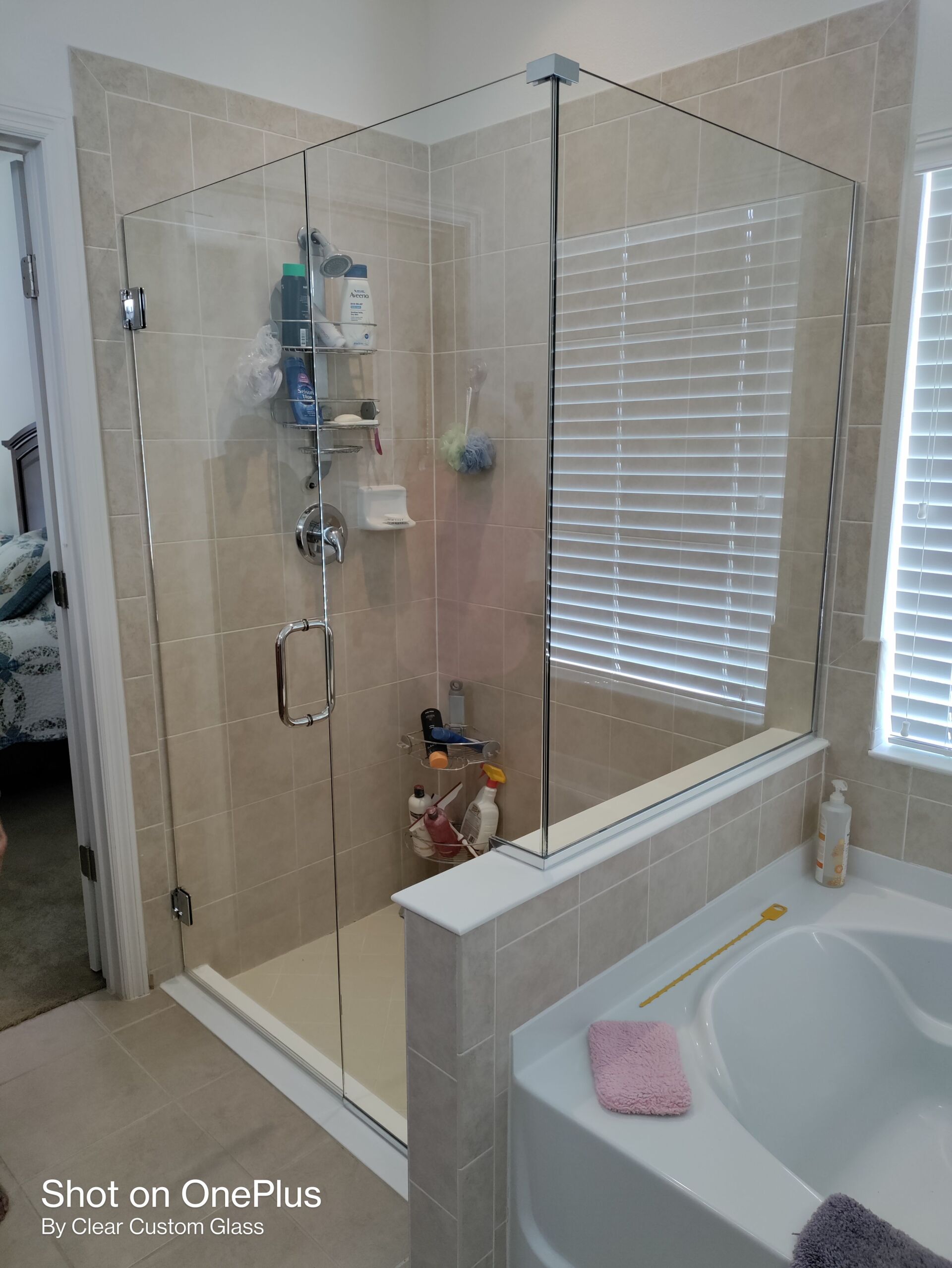 Glass shower enclosure with beige tile walls, a tub in the corner, and a window.
