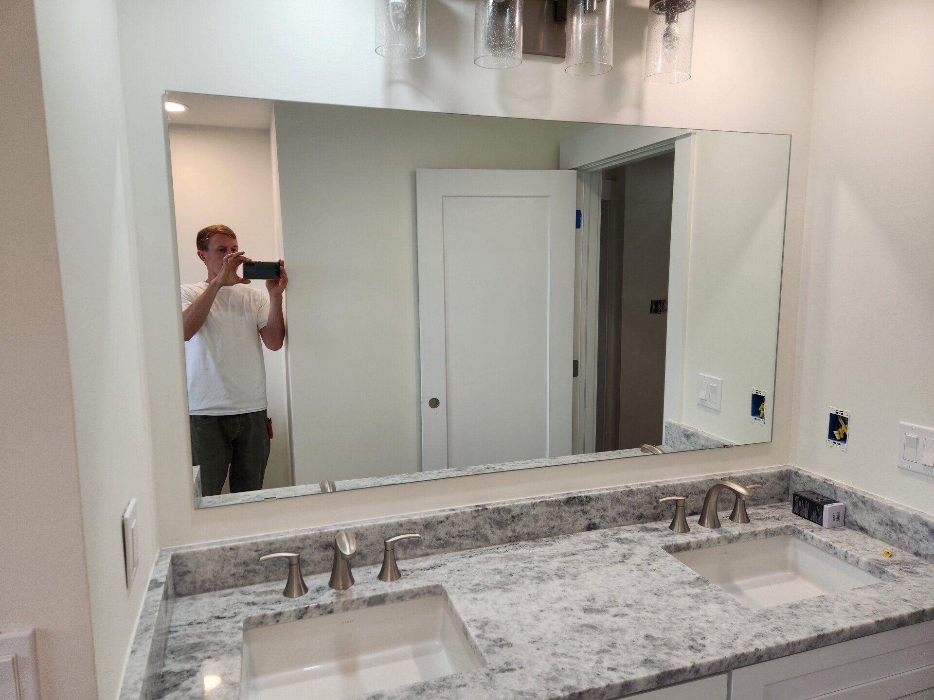 Person taking a photo in a bathroom mirror above a double sink, white cabinets, and light fixtures.