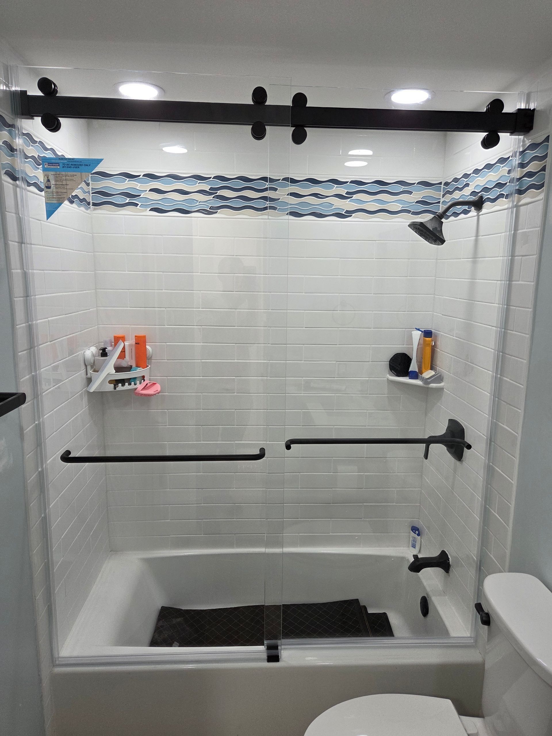 Bathroom with a shower over a bathtub, glass doors, and black hardware. White and blue tile accent the top.
