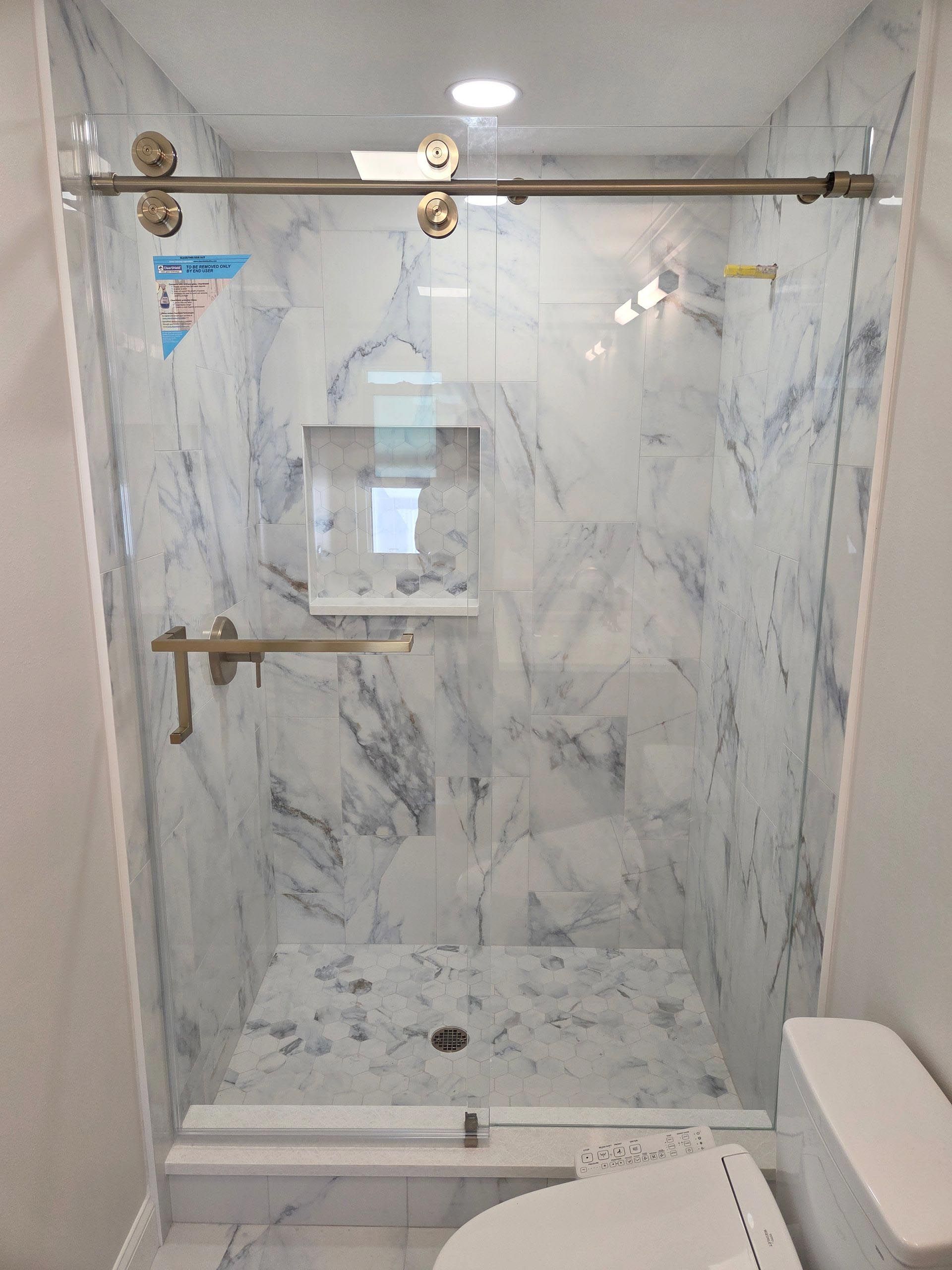 Marble-tiled walk-in shower with glass doors, brass grab bars, and wall-mounted controls