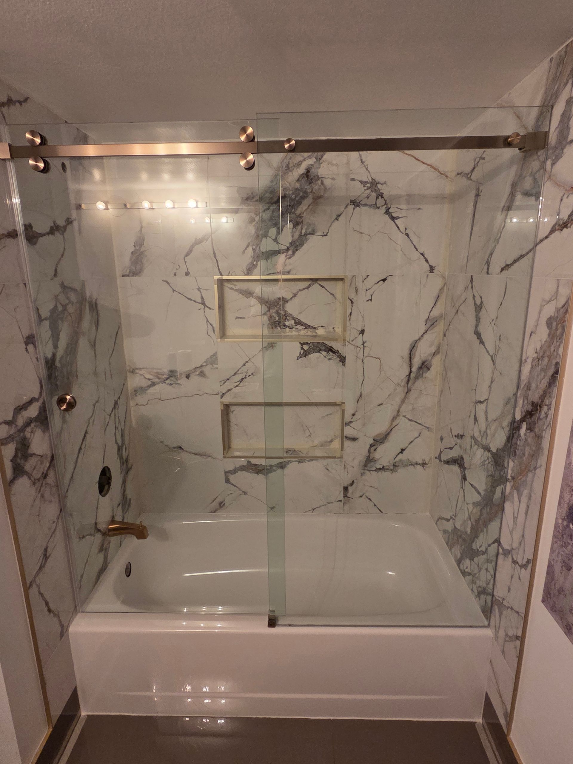Modern shower stall with marble-pattern tile, glass doors, built-in niche, and white bathtub.