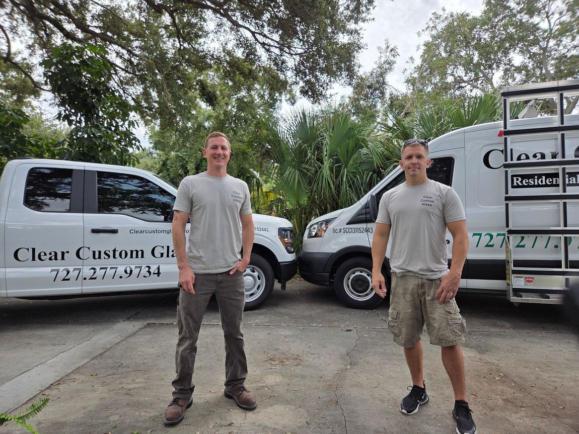 Two men standing by work trucks, wearing gray shirts, in a parking lot, by a tree, for Clear Custom Glass.