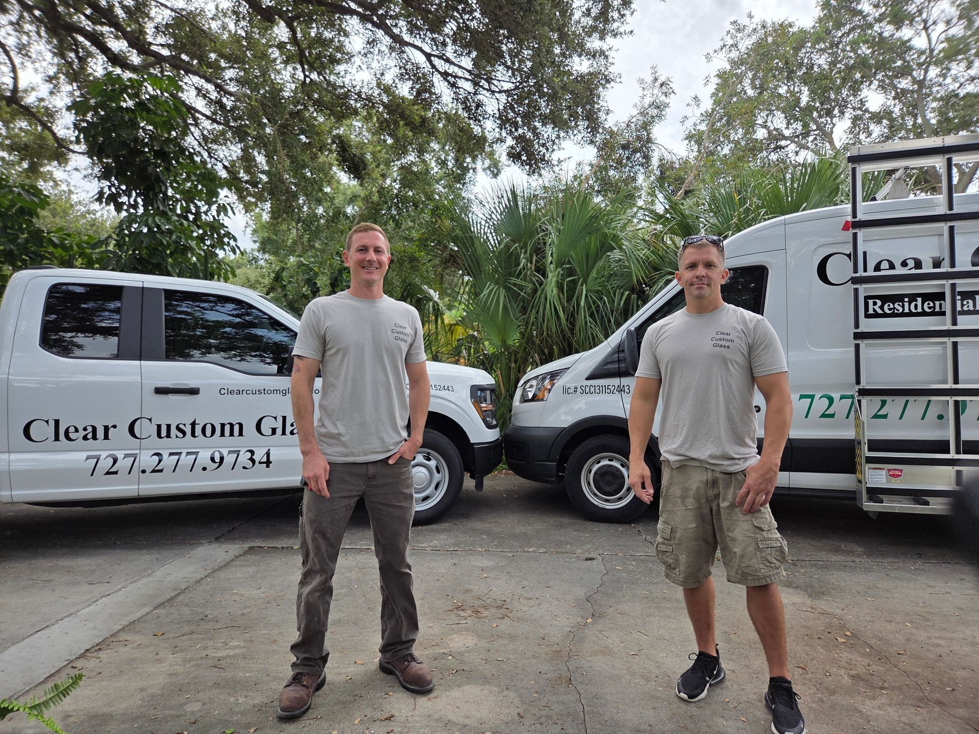 Two men standing by work trucks, wearing gray shirts, in a parking lot, by a tree, for Clear Custom Glass.