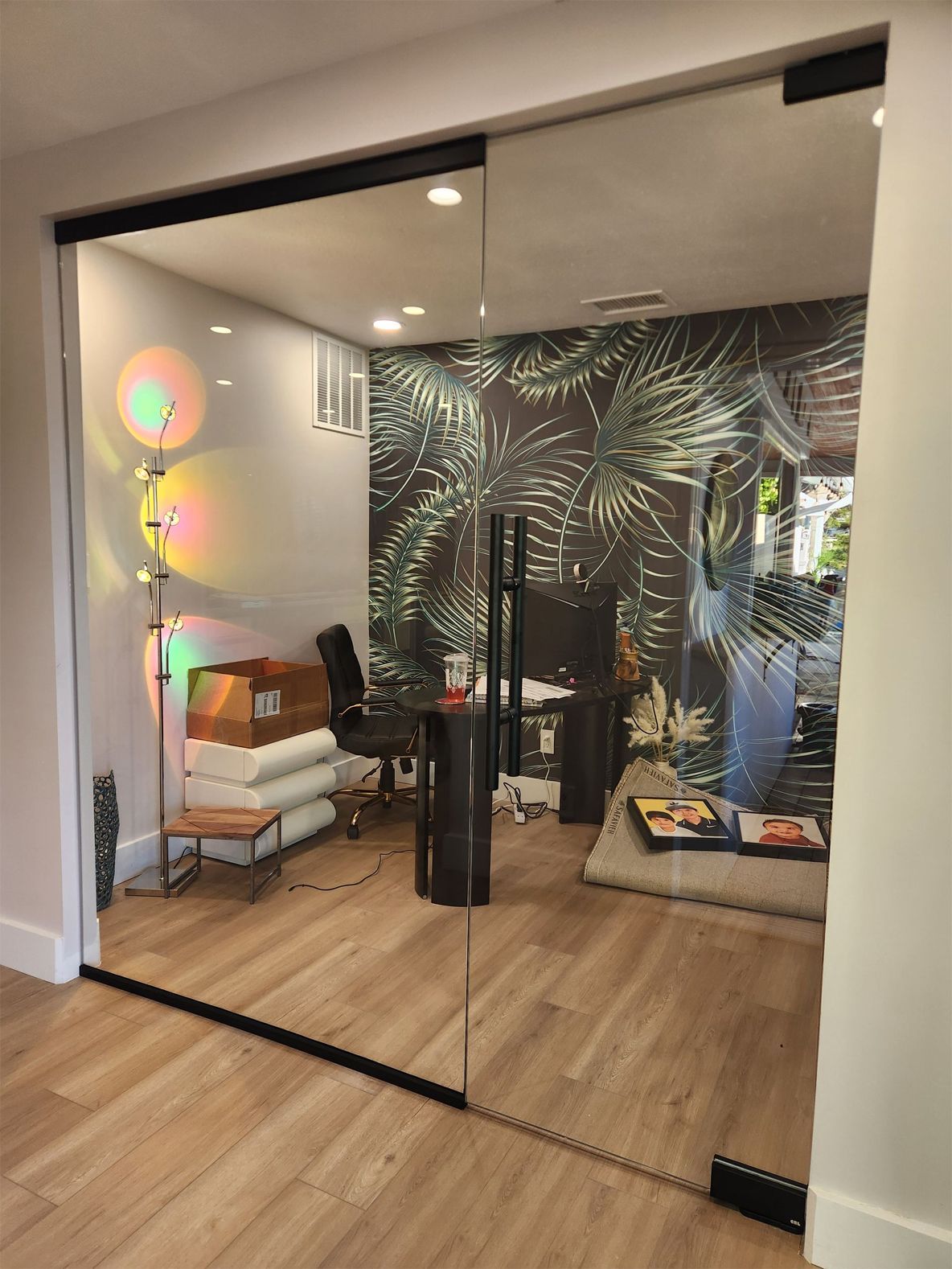 Glass-walled home office with desk, chair, and decorative palm-print wallpaper. Hardwood floor.