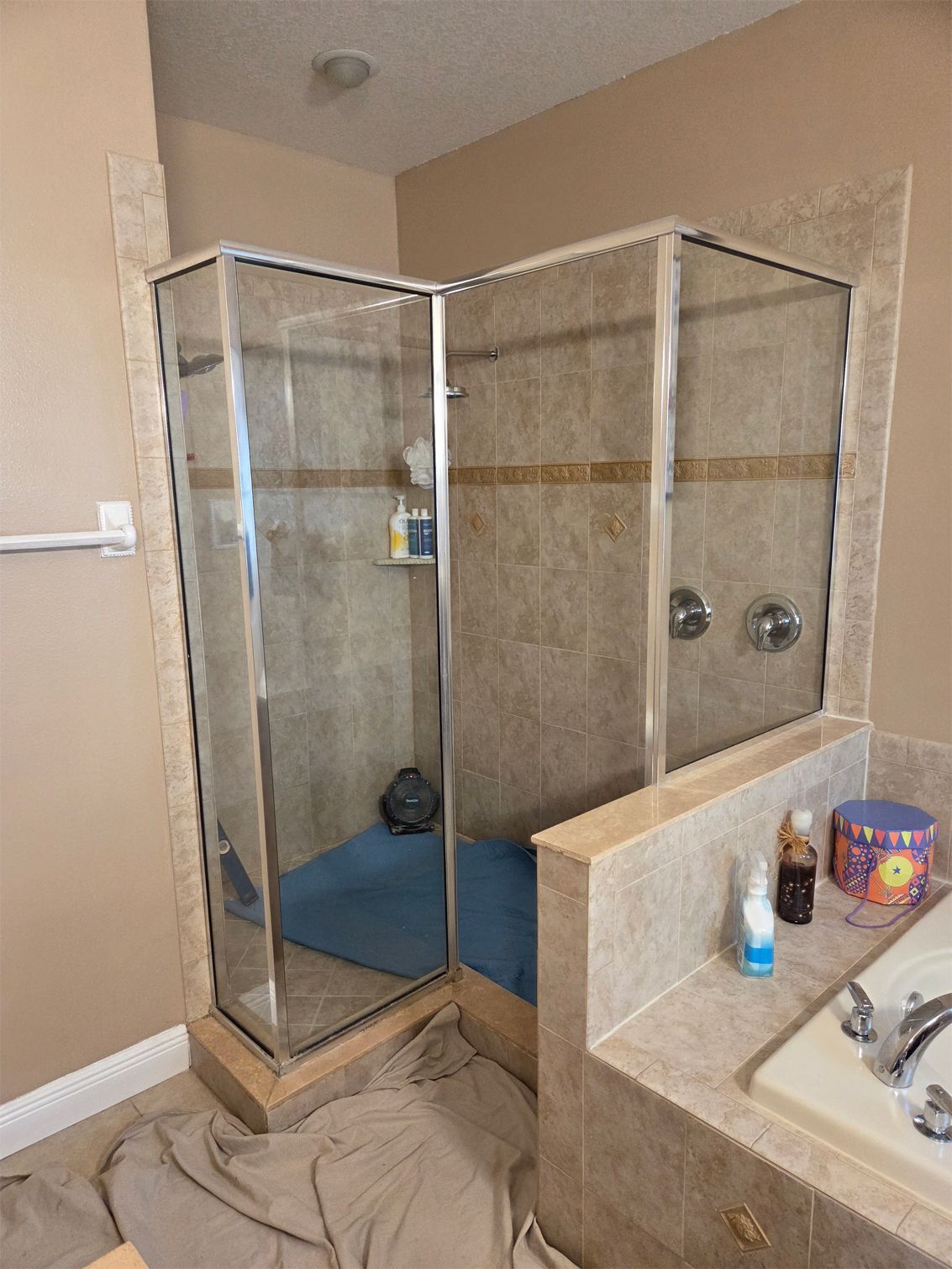 Corner shower with glass doors, beige tile, chrome fixtures, and a blue mat.