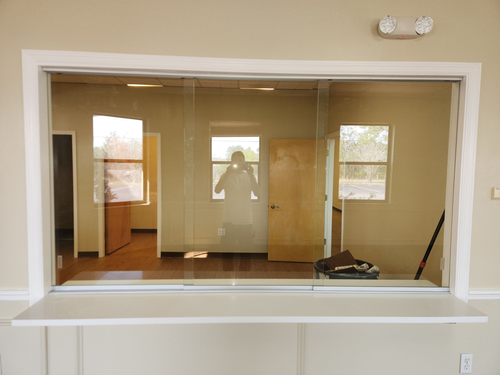 Interior view through a large service window with a person's reflection. Cream-colored walls, hardwood floors, and an open interior.