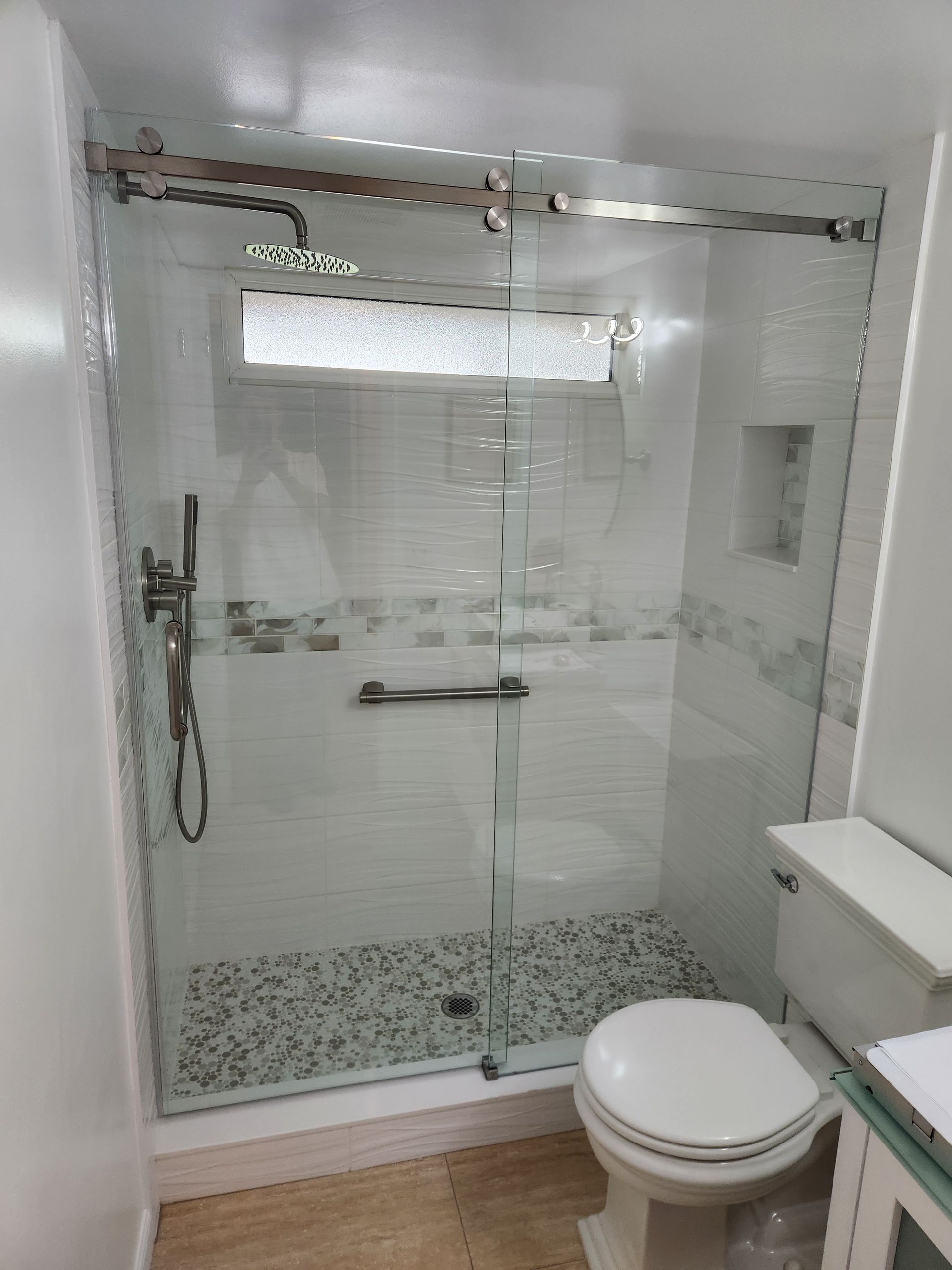 Shower with glass door, white tile, mosaic floor, grab bar, toilet, and neutral tones.