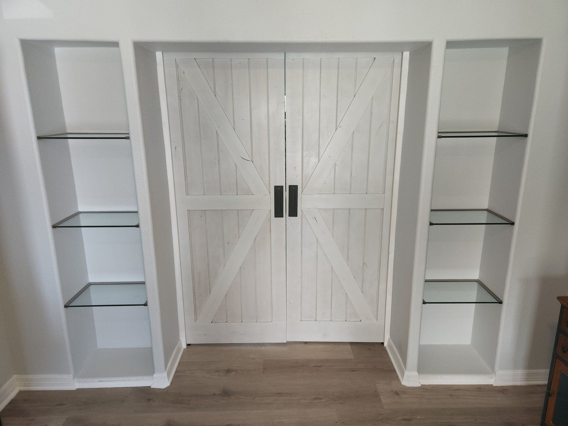 White built-in shelves flank a white barn door. Glass shelves hold items. Wood flooring.