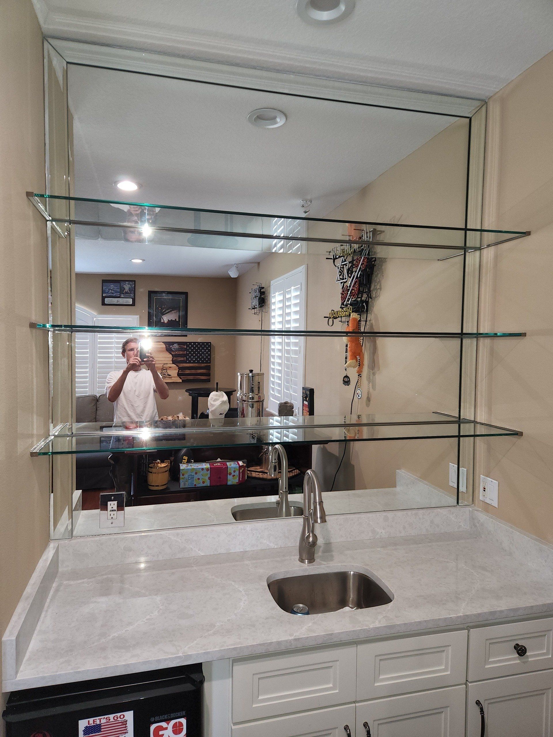A bar area with a sink, mirror, glass shelves, and a person reflected in the mirror.
