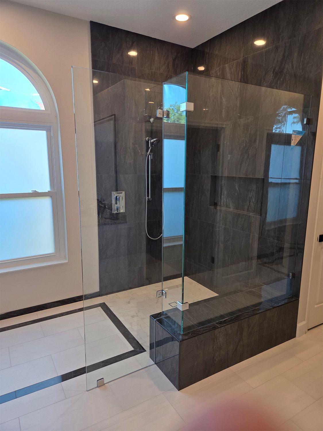 Modern bathroom with glass shower, dark tiled walls, and a frosted arched window.
