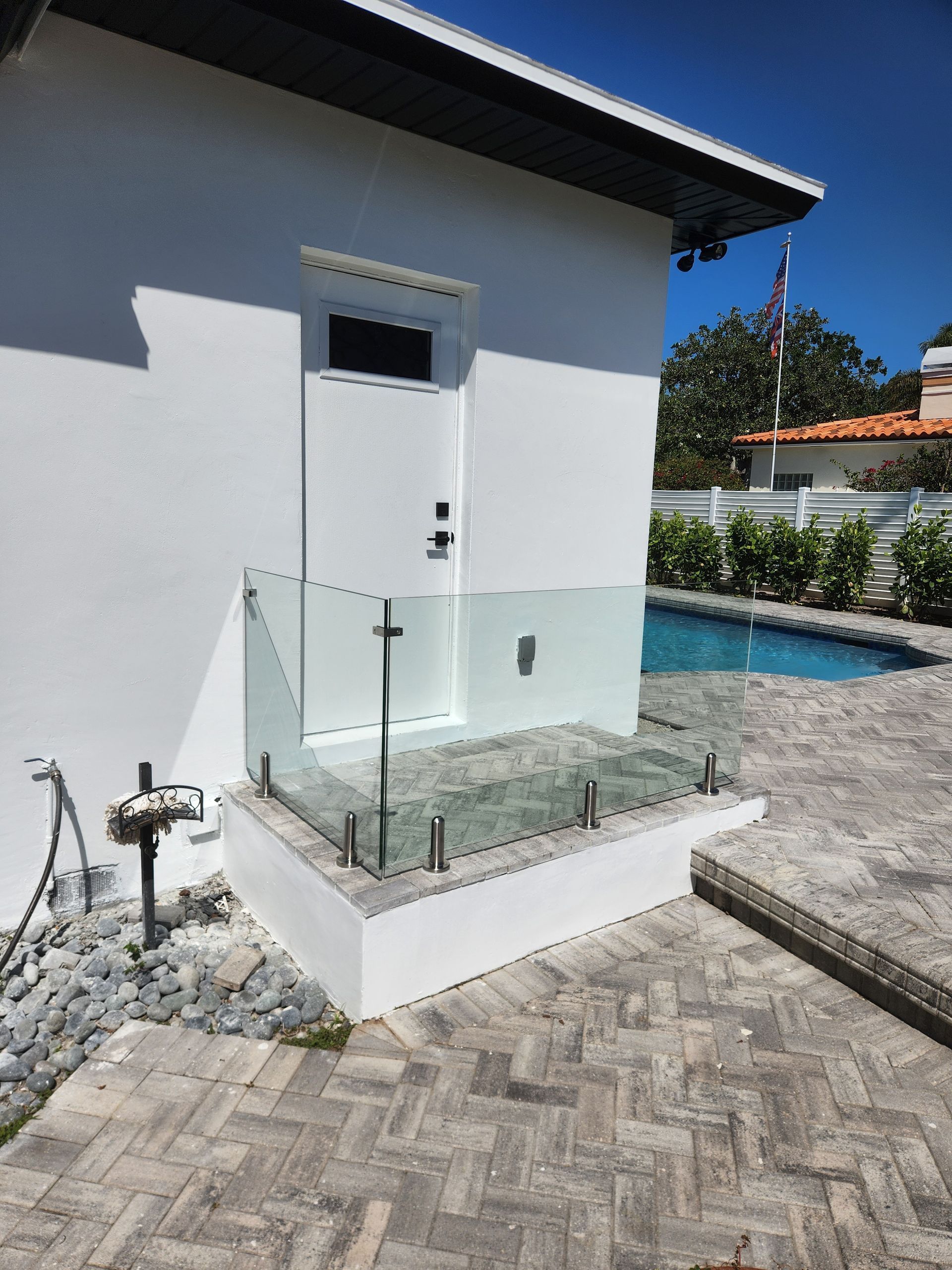 White building with glass railing, door, and pool in the background. Paved patio area.