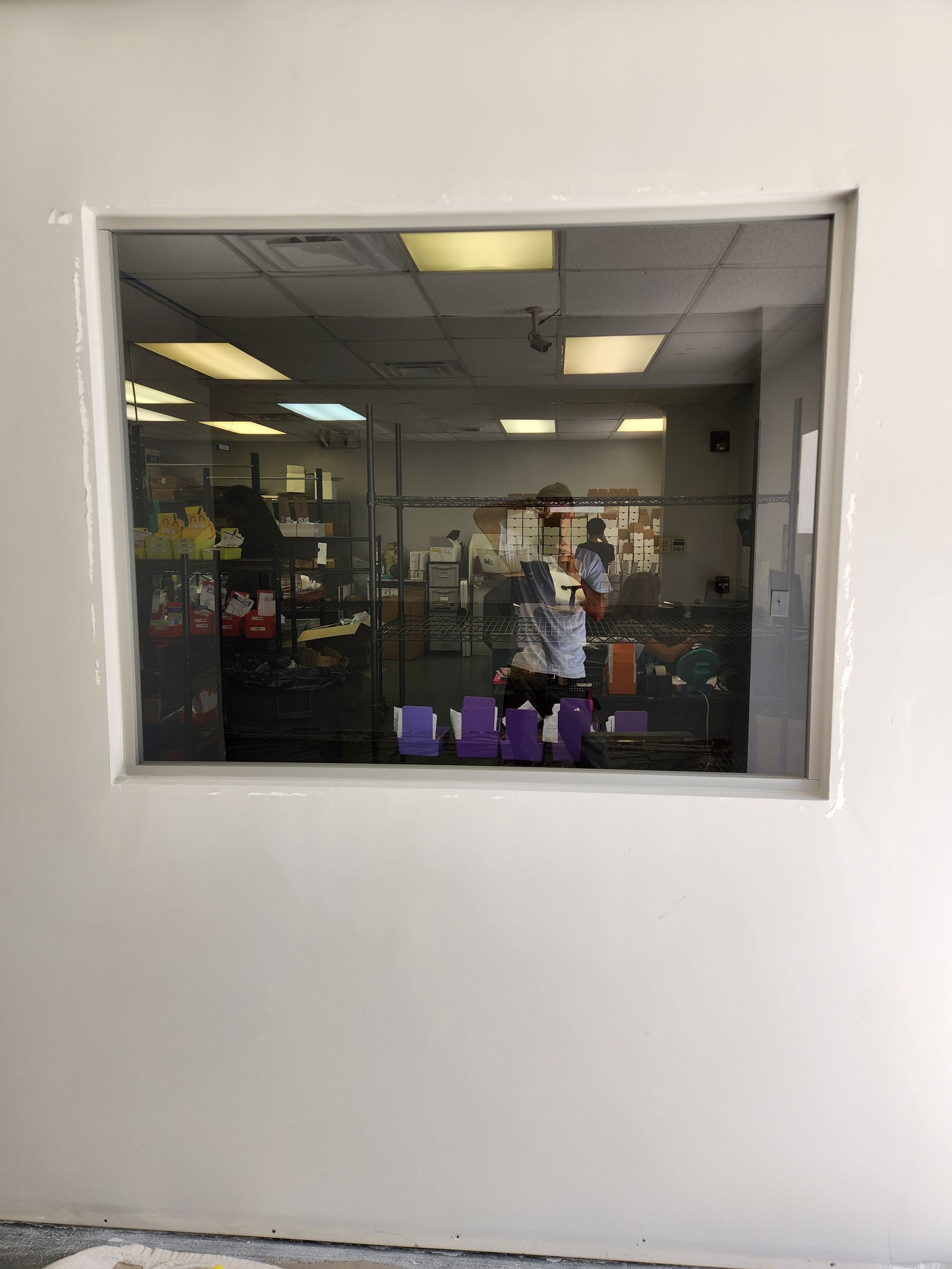 Window in a white wall. Inside the room are shelves, a person, and purple boxes.